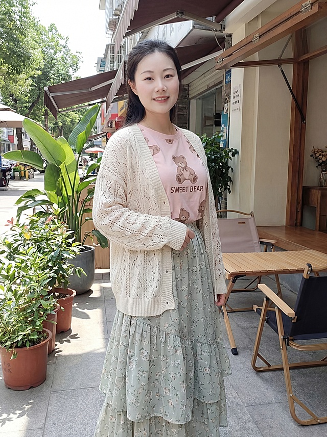 和上传人物保持一致性，葫芦形S身材，上围挺拔，沙漏型身材，傲人身材,- 外搭：米白色的针织开衫，采用镂空编织纹理，版型宽松休闲，增加了穿搭的温柔感。- 内搭：浅粉色的短袖T恤，印有棕色小熊图案及英文装饰，风格可爱俏皮。- 下装：浅灰绿色的长款碎花半身裙，裙摆带有多层设计，花朵图案清新淡雅，整体呈现出温柔甜美的日常穿搭风格