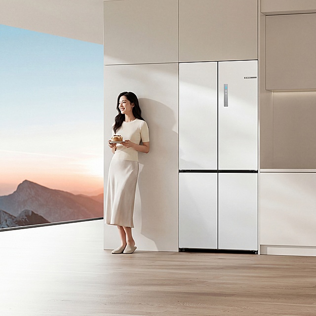 在冰箱左侧增加以为30岁左右的亚洲女性，微笑看着窗外，温柔优雅大气纤瘦女性，画淡妆，穿米白色修身T恤，浅色丝质半身裙，穿包头拖鞋，黑色微卷长发，背轻靠在冰箱边上的墙上，手上拿着蛋糕