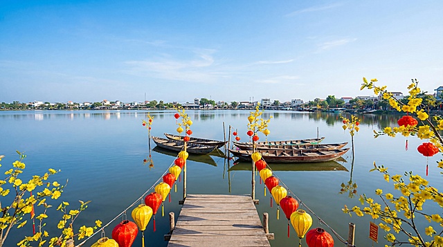 Mở rộng khung hình về phía trước và hai bên, thêm một bến chờ bằng gỗ đơn giản phía trước mặt hồ. Loại bỏ hoàn toàn con người, không xuất hiện người trên thuyền hay trên bến. Giảm chi tiết rườm rà, giữ bố cục gọn gàng và thoáng. Làm mặt hồ rộng hơn, phẳng và yên tĩnh. Mở rộng bầu trời cao và rộng, mây trắng nhẹ, ánh sáng ban ngày trong trẻo. Tạo không khí Tết miền Tây Việt Nam với sắc đỏ – vàng nhẹ nhàng, hoa mai và đèn lồng tre trang trí vừa phải. Phong cách ảnh chụp chân thực, màu sắc tự nhiên, không hoạt hình, không tranh vẽ.