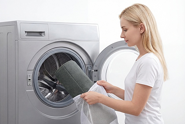 图上的记忆棉地毯是我的产品，请使用我的产品帮我生成亚马逊A+场景图，1940:1200比例，超高清：
主体为一位金发白人女性，身穿简约白色短袖，正侧身站在银灰色滚筒洗衣机前，双手将卷好的深灰色地垫装入白色网眼洗衣袋。洗衣机门呈打开状态，舱内细节清晰可见。
画面采用中景构图，人物动作自然舒展，焦点集中在手部与洗衣袋的互动上。整体色调以柔和的浅白、银灰为主，辅以洗衣袋的纯白与地毯的深灰，营造出干净清爽的视觉感受。
背景为纯净的白色墙面，无多余杂物，突出生活化场景的简洁质感。要求是高清写实电商场景图，细节清晰，无文字，纯产品展示，聚焦视觉卖点，排版简洁干净。