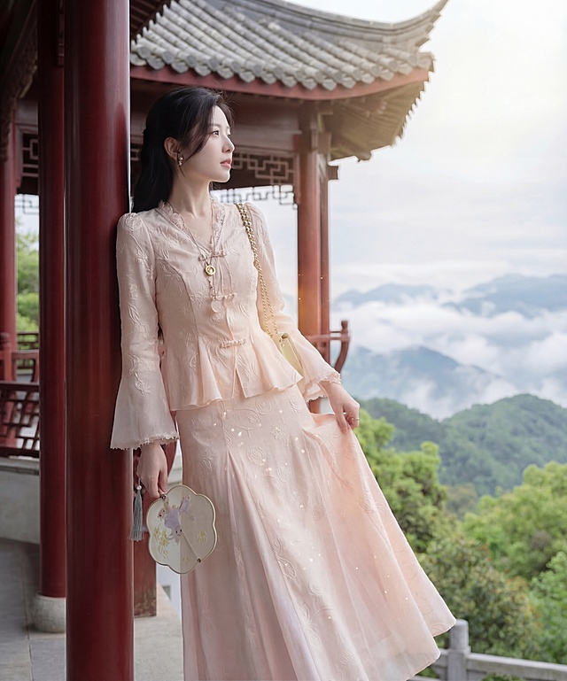 一位女士身着烟粉色提花中式套裙，上衣是V领蕾丝花边盘扣收腰裙摆设计，下装是同面料同色系长款半身裙，女士侧身倚在云台山木质观景廊的朱红立柱上，一手轻提裙摆，一手握着绣着玉兔衔花的团扇，肩上斜挎着米白色链条包包，目光望向远方云海，姿态松弛端庄。上午十点的柔光穿过山间薄雾斜洒在肩头，把烟粉色衣料晕染得暖而朦胧，提花纹路在光线下忽明忽暗像山野跳动的光斑。背景是朱红立柱与青灰瓦当的木质观景廊，廊外是层叠青山与漫山新绿，远处云雾缠绕山尖，中式雅致与山野辽阔融合，风拂过发梢与衣摆，整体氛围清冷遗世，如从山水画卷走出的林间仕女。