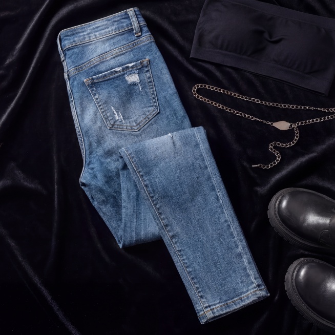 保持图中蓝色牛仔裤的颜色、纹理、版型、磨损细节完全不变，将其调整为背面朝上摆放的造型，展示牛仔裤背面设计，背景为黑色丝绒布艺，搭配黑色抹胸、金属腰链、厚底靴道具不遮挡裤身，影棚级逆光+顶光柔光补光，裤预览效果