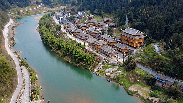 将沿河小镇改造为水族风情文化旅游景观，房屋改为传统水族风格风貌：1.采用干栏式木楼结构，底层架空用于饲养牲畜和堆放农具，上层为居住区域；2.屋顶改为重檐歇山式，覆盖青瓦或杉木皮，屋檐翘起如飞鸟展翅；3.建筑采用松木、杉木为主要建材，呈现自然原木色；4.柱脚采用鱼尾状榫卯连接，增强结构稳定性；5.门前添加水书对联装饰；6.沿河景观增加水族文化元素，如水族图腾、鱼形装饰；7.房屋布局保持'左-中-右'三开间形式，中间为堂屋；8.沿河种植翠竹、花树，形成'青山绿水环绕，花果飘香'的生态景观；9.保留原有道路和部分现代设施，但外观统一为水族风格；10.整体色调以自然木色、青灰色为主，营造古朴和谐氛围。