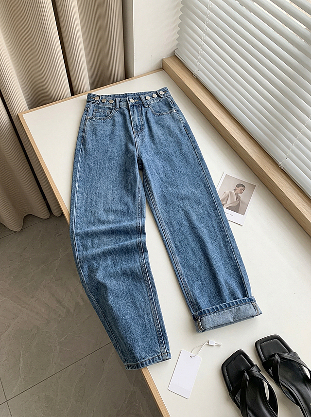 45°斜俯拍电商服装平铺摄影，白色窗台桌面，右侧白色竖条百叶窗，左侧竖纹布艺窗帘，浅灰色瓷砖地面，搭配一张人物小卡片、白色吊牌、一双黑色方头凉鞋，柔和漫射自然光，干净明亮氛围感，高腰蓝色牛仔裤（裤腰多扣设计，牛仔布纹理与参考图完全一致，保持主体物理结构不变），裤腰朝上放置于桌面左上角，左腿裤脚自然堆叠褶皱，右腿裤脚翻边，高清质感，真实细腻，全屋漫射柔光，低对比度，画面明亮通透，莫兰迪奶油色系，前中后景层次分明，三分法视觉重心