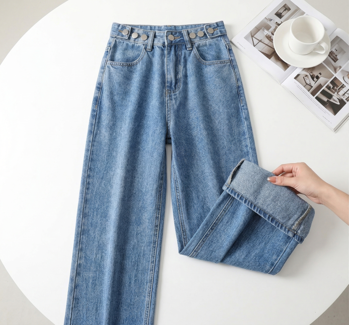正俯拍电商服装细节展示摄影，主体为高腰浅蓝色牛仔裤，保持产品主体物理结构不变，保留裤子腰头纽扣和牛仔布料纹理，纯白色平整桌面背景，右上角搭配带白色杯碟的杂志页，右侧有一只手轻托裤身掀起面料以展示牛仔肌预览效果