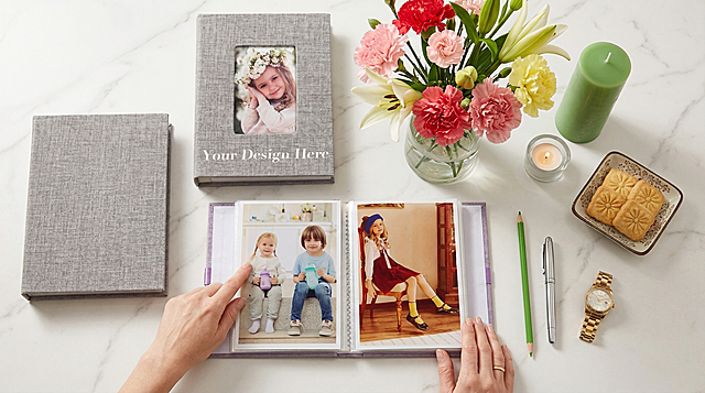 俯拍视角，保持原图相簿原有结构不变，封面文字保持原样。画面中宁静欧美女性左手轻指着摊开相册内页，右手按压在相册角落，干净白色大理石台面放置产品。旁边放置另一本相同相册，透明玻璃花瓶中插着粉、红、黄三色康乃馨与白色百合组成的鲜艳花束，旁侧立着绿色柱状蜡烛与点燃的小茶蜡，还有绿色铅笔、银色钢笔、金色腕表、方形花纹糕点。柔和自然光洒落，暖调舒缓配色，点缀柔粉、艳红、浅黄与清新绿意，超写实摄影质感，清晰对焦，光影柔和，Ins风审美，整体氛围温馨自然，商业高调摄影，柔和漫射光，前中后景层次分明。负向提示词：亚洲面孔、韩流风格、苍白皮肤、柔焦模糊、背景虚化过度、平光、动漫风、可爱道具、多余的盖子、缺失的部件、悬浮的物体、变形的手柄、摄影灯、柔光箱、三脚架、水印、文字、昏暗光线、死黑阴影、畸变透视、比例失调