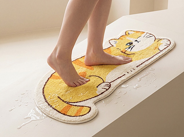 橘黄色猫咪造型毛绒地毯，保持产品本身的外形图案和材质纹理不变，严禁扭曲变形。将地毯放置紧贴在45度的防滑坡度中间，一双东亚美女的光脚双脚前后踩在地毯上，双脚深深陷入柔软蓬松的地毯面料中，清晰展示面料的柔软质感，斜坡周边分布着零星透明水渍，展示地毯防滑吸水的产品特性。
整体为浅色系干净简约背景，保持产品主体清晰突出。
采用柔和漫射柔光，画面明亮干净，低对比度，营造温馨家居种草氛围感。
商业产品摄影，4K高清分辨率，清晰展现材质纹理。