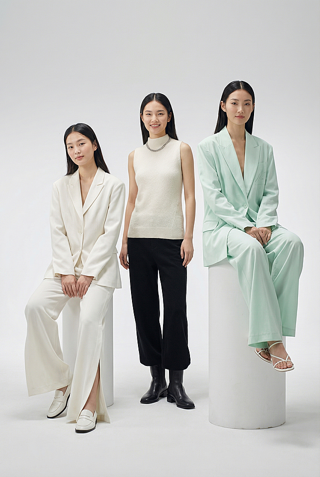 将参考图中的假人模特替换为三位真实亚洲模特，模特穿着和参考图完全一致的服装：左侧模特穿米白色宽松西装搭配侧边开叉阔腿长裤和白色乐福鞋，保持坐姿，神态柔和自然；中间模特穿米白色无袖针织上衣搭配黑色阔腿长裤，佩戴银色链条项链，穿黑色皮面短靴，保持站姿，温柔浅笑；右侧模特穿薄荷绿宽松西装套装搭配同色系阔腿长裤和白色细带凉鞋，坐在白色圆柱台座上，姿态放松优雅。整体背景为干净极简的白色影棚，使用柔和漫射光，打造高端时尚大片风格，要求8K分辨率，超精细细节，清晰对焦，自然真实的皮肤质感，专业时尚摄影，保持参考图中每个人物的原始姿态不变。