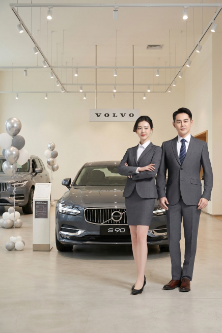 重新生成背景为明亮通透的沃尔沃汽车展厅，北欧简约风格，浅色调墙面与地面，轨道灯柔和照明。正中央展示深灰色沃尔沃S90轿车，突出直瀑式镀铬格栅与沃尔沃铁标，天花板悬挂 "VOLVO" 品牌标识，左侧有气预览效果