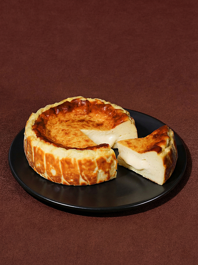 将原图黑色盘子上的蛋糕原位替换为图1(参考图)中的切开的流心巴斯克芝士蛋糕。
用图1中蛋糕的真实物理结构完全覆盖该区域，打破原图旧蛋糕的轮廓束缚，保持原产品(图1)的真实比例，严禁挤压或变形。新产品需完美继承原图中旧蛋糕的摆放角度、镜头高度与透视消失点关系，继承旧蛋糕的底部接触点，严禁漂浮或位置偏移，使其与背景空间完美融合。仅替换蛋糕主体，保留黑色盘子和背景环境不变。
重置该区域光影，赋予自然接触阴影，完美消除新产品边缘白边，画面自然真实，严格锁定宽高比为 3:4，4K分辨率。