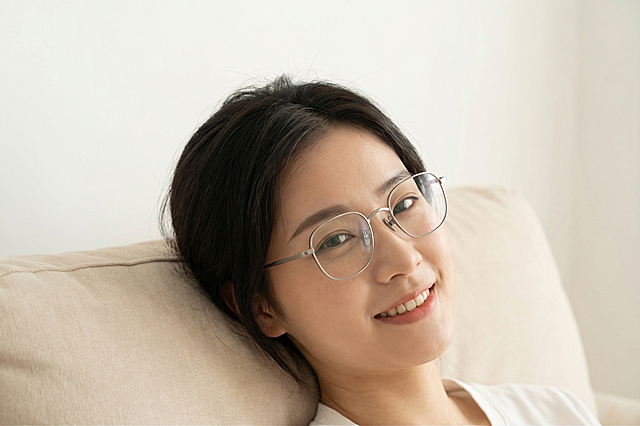 保留这副银色金属细框眼镜的款式、颜色和材质完全不变，将其佩戴在一位东亚女性模特脸上，模特表情自然惬意，姿态松弛倚靠，背景简洁纯色，保持整体画面明亮干净，风格为国内电商种草氛围，严禁产品结构变形扭曲