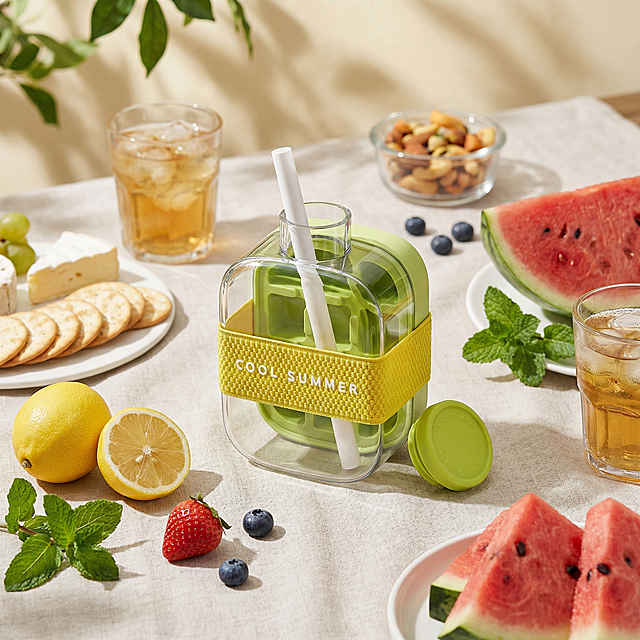 cozy outdoor picnic lifestyle image, this portable ice-making cup placed as the visual hero on a light picnic cloth, surrounded by fresh fruit, cold drinks, and summer snacks. Soft sunshine filtering through, gentle natural shadows, relaxed and happy atmosphere. Keep the product's transparent outer shell, green inner ice mold, white straw, yellow textured elastic band with text "COOL SUMMER" and green lid original structure unchanged. Looks refreshing, useful and emotionally appealing. Premium lifestyle product photography, modern summer drink accessory, high-end product photography, ultra realistic, clear transparent material, soft natural lighting, realistic details, premium lifestyle aesthetic. Keep 1:1 square ratio.