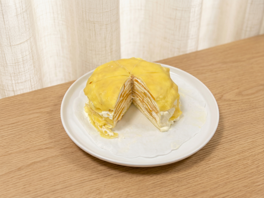 保持千层蛋糕（切开露出多层结构）和白色盘子的物理结构绝对不变，严禁变形。更换为温馨治愈的美食场景：置于浅橡木纹桌面，背景为米白亚麻布帘，柔和漫射光，低对比度莫兰迪暖调，画面干净明亮，商业种草风，4K高预览效果