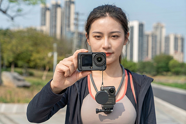 将图片中的原人物替换为一位美女，她穿着运动服，保持原图片中手的姿势，握住运动相机。保留原图片中的黑色运动相机、镜头盖和挂绳这些元素，背景保持原来的户外城市树木街景不变