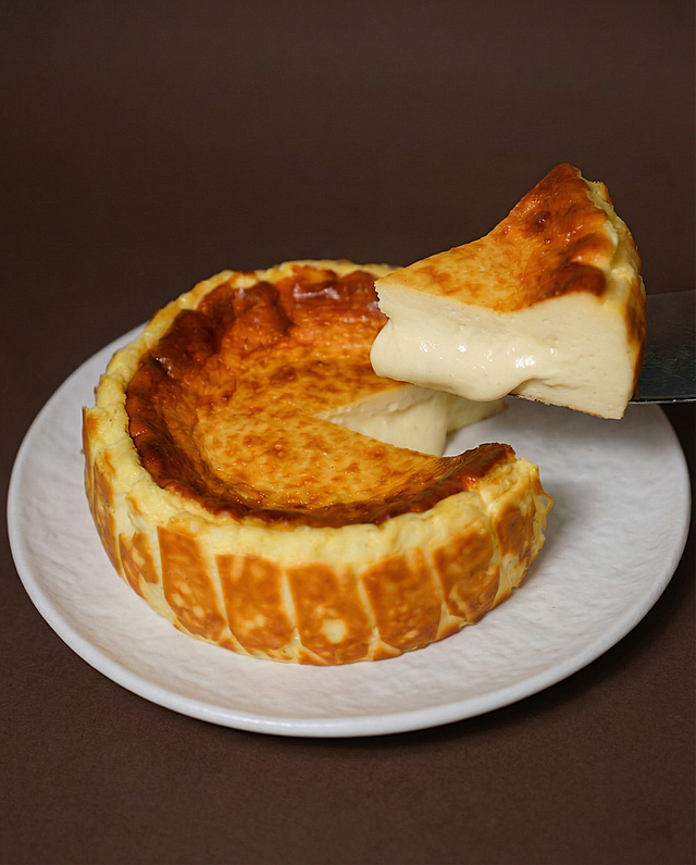 将图1的巴斯克芝士蛋糕整体迁移放进图2的场景中，保证蛋糕物理细节完全不变。
精准匹配图2场景的透视角度，实现空间尺度物理融合，蛋糕自然嵌入场景。
重置蛋糕光影，增强光照一致性，赋予蛋糕符合环境的自然光影及真实接触阴影。
蛋糕色温亮度与深棕色背景环境完全协调，保持蛋糕烘焙质感。
消除视觉接缝，画面自然真实，保持原背景色调，强化商业摄影质感。
保持原图比例，4K分辨率。
负向提示词：抠图白边,阴影方向错误,光影不匹配,透视错误,空间比例失调,巨物感,微缩模型感,悬空,违背物理常识,贴纸感,视觉接缝,边缘生硬,变形,扭曲,画面被裁剪,比例改变,背景被篡改