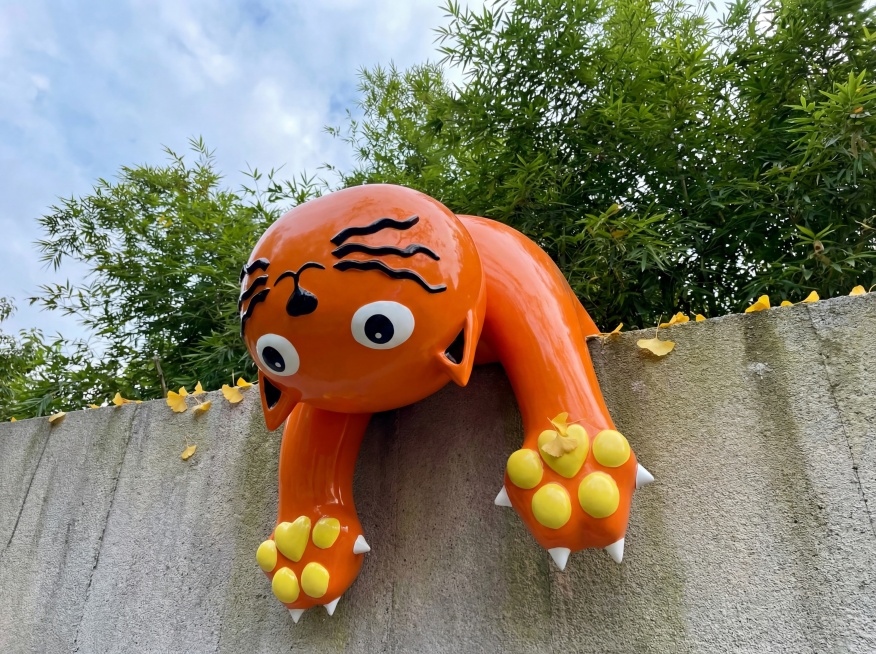 产品是亮橙色玻璃钢小猫穿墙雕塑，黑色条纹，黄色爪垫，白色尖爪，保持这个物理结构和外观绝对不变，严禁扭曲变形。设计在庭院灰色文化石围墙上，背景是茂密的绿色竹子和蓝天，自然户外氛围。阴天柔和自然光，低对比预览效果