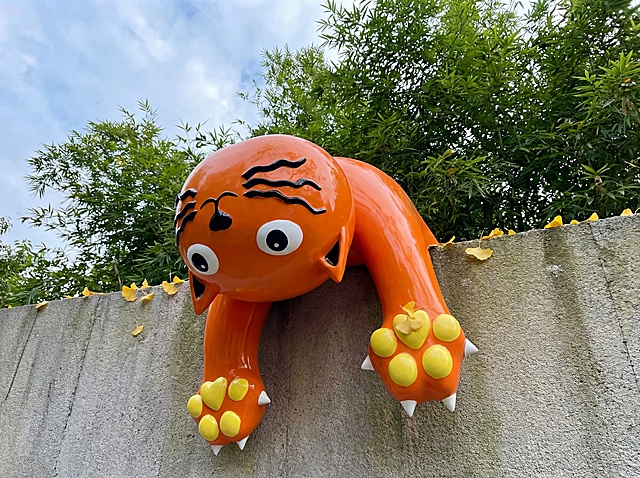 产品是亮橙色玻璃钢小猫穿墙雕塑，黑色条纹，黄色爪垫，白色尖爪，保持这个物理结构和外观绝对不变，严禁扭曲变形。设计在庭院灰色文化石围墙上，背景是茂密的绿色竹子和蓝天，自然户外氛围。阴天柔和自然光，低对比度，整体色调偏清新自然。仰拍视角，让雕塑和天空绿植形成层次。周围点缀几片掉落的银杏叶，自然生机感。商业级4K，画面通透，保持原图比例。西方面孔,金发,深眼窝,雀斑,杂乱房间,死黑阴影,硬光,乡村风,高对比度,欧美妆容,变形,扭曲,悬空,比例失调