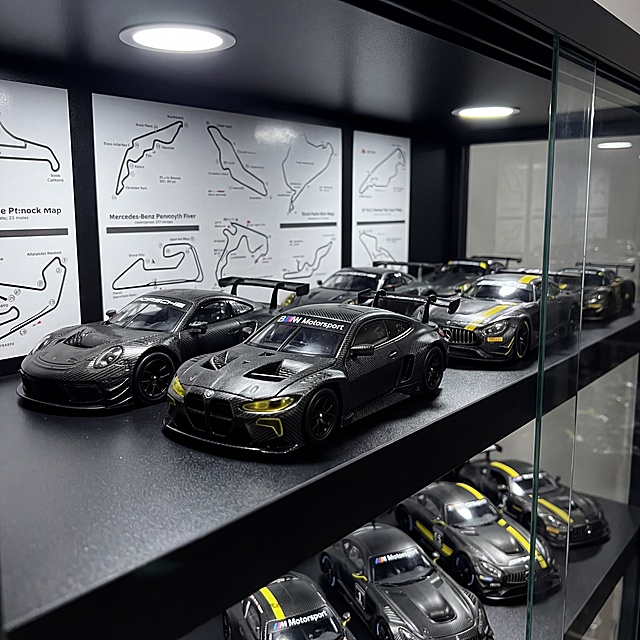 保持原BMW赛车模型的黑色碳纤维纹理、黄色灯组细节、BMW Motorsport标识以及整体物理结构绝对不变，将模型放置在黑色哑光分层玻璃展示柜的中层C位，周围环绕同系列保时捷、奔驰赛车模型，所有模型按赛道队形排列，本款BMW模型在前领航。展示柜顶部安装冷光射灯，重点打亮模型碳纤维纹理与黄色细节，柜内背景贴赛道地图贴纸，营造专业赛车收藏柜的氛围，商业摄影，4K超清，细节清晰，保持原比例。