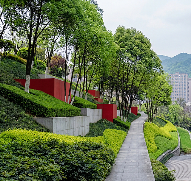 将第二张图片中的绿化山坡场景，按照第一张图片的风格进行渲染：保留原有构图和植被布局，但采用第一张图片的色彩调性、材质质感和整体氛围。第一张图片显示的是生物医药产业园的实景，有红绿相间的色块装饰、混凝土结构墙体、远处山林背景和阴天光线效果。要求保持第二张图中树木、灌木和小径的布局，但赋予其类似产业园的工业感与规整感，色彩更饱和，光影更均匀，体现现代园区风貌。