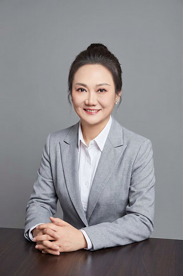 参考图中保留人物脸型、发型、年龄等特征生成教育嘉宾职场形象照,标准焦段50mm,f/4光圈,平视视角,人物居中,半身像,头肩比例协调。身穿浅灰色西装外套内搭白衬衫,着装正式干练,知性亲和。自然坐姿双手交叠放桌面,表情自然嘴角上扬。中度灰色纯色背景,光线均匀柔和,极简干净。32k高清画质,细节清晰,清透自然裸妆,肤色均匀,专业精致写实风格。去除多余元素，严格按照参考生成。