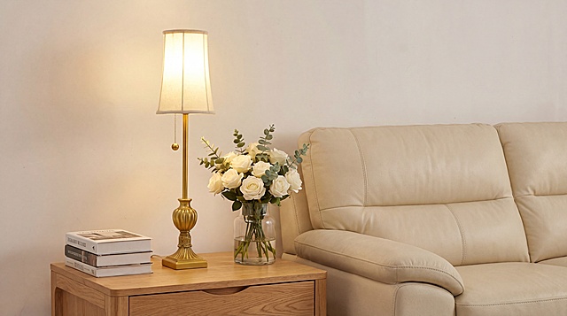 写实现代简约美式风温馨家居电商产品宣传图，暖调治愈的商业展示氛围，平视正面视角，左右构图，核心产品为台灯，台灯使用参考图一的产品以及角度，产品角度保持不变，台灯散发参考图二一样均匀的白色柔光；左侧台灯放置在浅原木色实木边几上，左侧边几台面上摆放精装书籍，台灯放中间，旁侧搭配透明玻璃花瓶，花瓶中插满白色玫瑰与尤加利叶，右侧背景为米白色软包皮质沙发