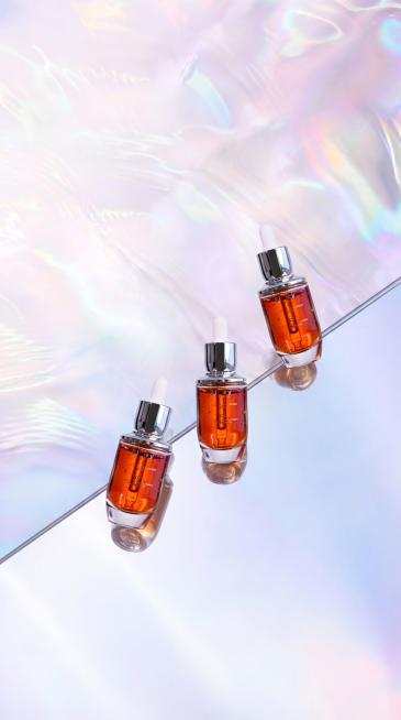 将原图镜面上斜对角排列的三个白色圆柱瓶子原位替换为图2(参考图)中的琥珀色透明玻璃瓶精华液，瓶子带有银色滴管式瓶盖，瓶身有文字标识。
用图2中精华液的真实物理结构完全覆盖该区域，打破原图旧瓶子的轮廓束预览效果