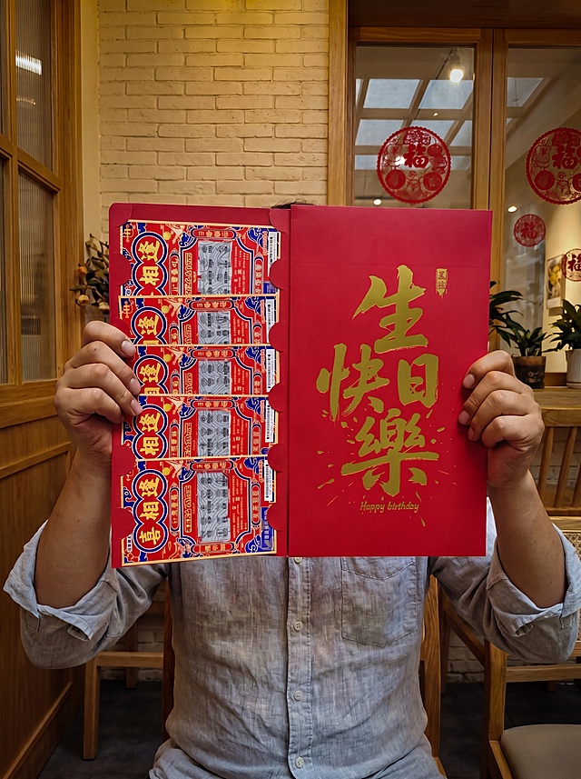 调整光影：将背景墙壁调暗，降低整体亮度，增强自然光和室内光的真实柔和感，让红包、人物与背景光影完全协调，无局部过曝和过暗；保留红包烫金细节的自然光泽，不破坏整体暗调和谐度。其余内容保持不变：人物穿着男性长袖夏装，双手对称持握超大展开式红包，面部被红包遮挡，温馨治愈的室内咖啡轻食店场景，暖黄色柔和光线，背景浅米色砖墙、木质窗框、玻璃窗红色圆形装饰，充满生日氛围感