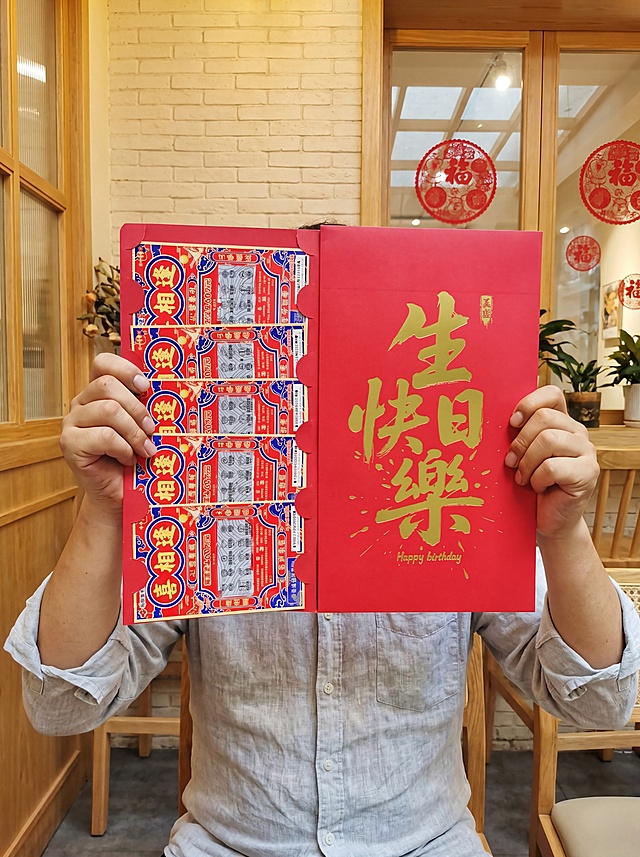 基于原图修改：将人物衣服更换为男性长袖夏装，其余内容保持不变。画面核心依旧为人物的双手，呈左右对称持握超大展开式红包的姿势：右手在画面左侧，稳稳捏住红包左页的边缘；左手在画面右侧，稳稳捏住红包右页的边缘，双手将红包完全展开、举在胸前，中间为完整的、适配超大红包的区域；手臂自然舒展，手指关节清晰，姿态放松自然，完全适配超大体积红包的托举需求，人物面部完全被红包遮挡，无露脸。保持温馨治愈的室内咖啡/轻食店场景，暖黄色柔和的室内自然光，背景可见浅米色砖墙、木质窗框、玻璃窗上的红色圆形装饰，整体氛围轻松日常，充满生活化的生日氛围感，无杂乱杂物干扰主体，保持红包上的原有金色字体“生日快樂 Happy birthday”和刮刮乐区域内容