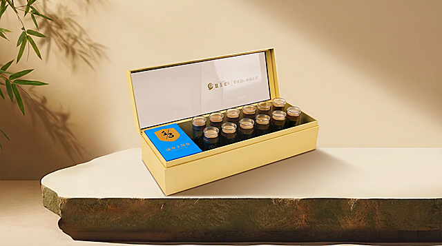 将原图石材台面上的旧产品组合替换为图1(参考图)中的打开的金色长条海参礼盒。
缩小产品尺寸，移除手提袋，增大画面留白，用图1中物体的真实物理结构完全覆盖该区域，打破原图旧物的轮廓束缚，保持原产品(图1)的真实比例，严禁挤压或变形。新产品需完美继承原图的摆放角度、镜头高度与透视消失点关系，严禁改变透视，继承底部接触点，严禁漂浮或位置偏移，使其与背景空间完美融合。仅替换该物体，保持原图其他环境布局绝对不变。
重置该区域光影，赋予自然接触阴影，完美消除新产品边缘白边，画面自然真实，严格锁定宽高比为 2:1，4K分辨率。