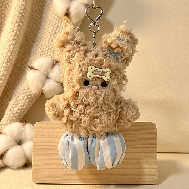 主体是浅卡其色卷毛毛绒兔子挂件，头部带彩色波点骨头发饰，穿蓝白条纹短裤，顶部带有心形金属挂扣。保持物理结构不变，严禁扭曲拉伸改变产品形态。
温馨室内软装场景，暖棕奶油色调，搭配柔和棉絮质感装饰。
柔和侧逆光。【产品受光面泛起暖奶黄色，暗部及边缘映射浅棕米色的环境光晕。绝对禁止纯白高光，彻底物理融合】。
画面居中特写摆放。自然柔和光照，产品悬挂展示。
微调拍摄视角。搭配原木立牌（替换帆布包/线圈本），背景用米色磨砂台面重构细节，消除品牌文字。
严格锁定宽高比为 1:1。4K分辨率