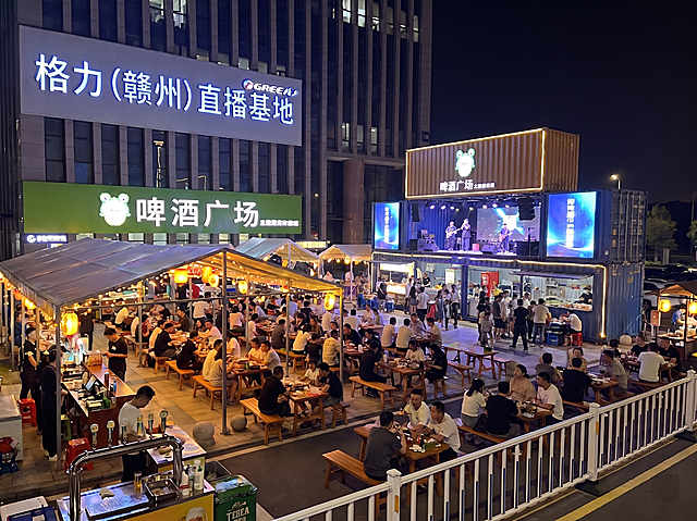 将现有啤酒广场的独立舞台替换为集装箱结构，集装箱二楼作为舞台，保留原图中格力赣州直播基地招牌、啤酒广场绿色招牌、用餐座位和人群，保持夜晚夜景氛围，不需要单独搭建舞台，保留原有灯光和热闹氛围