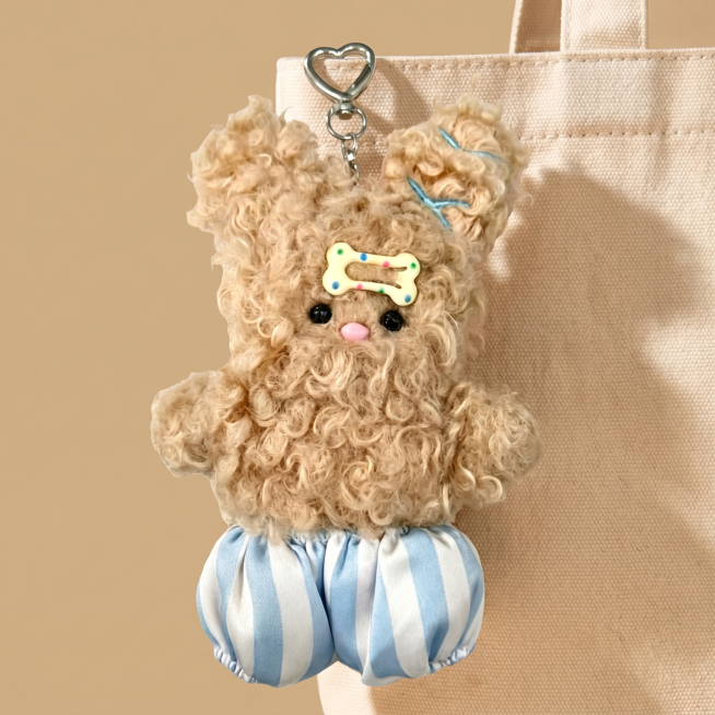 浅卡其色卷毛毛绒小兔挂件，金属心形挂扣，头上装饰带彩色波点的奶黄色骨头发夹，搭配蓝白竖条纹灯笼短裤。保持产品原有外观、形状、颜色和材质不改变，严禁扭曲拉伸。
挂在米色帆布包包提手处的展示场景，暖调浅卡预览效果