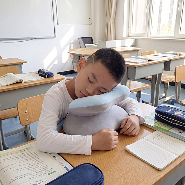 保持图中小男孩样貌、发型和午睡姿势不变，将背景场景更换为明亮的教室课桌上，把人物衣服的短袖换成白色长袖，午睡枕保持蓝色带胸垫的外形不变，真实自然的实拍效果