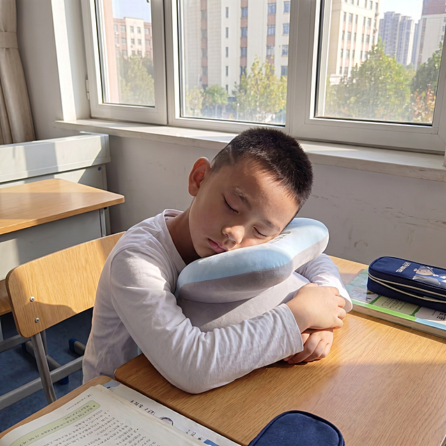 保持小男孩样貌、发型不变，调整姿势为手臂环绕抱住午睡枕，保持蓝色带胸垫午睡枕不变，移动位置到教室窗户边的课桌上，阳光透过窗户，真实自然的实拍效果