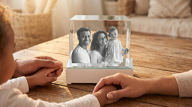 保持原图中玻璃展示盒摆件的外形不变，将摆件放在木质桌面上，桌面伸出年轻爸爸、妈妈和三岁孩子的三只手牵在一起，三只手将摆件包围在中间，营造出温馨温暖的氛围，整体场景美观自然