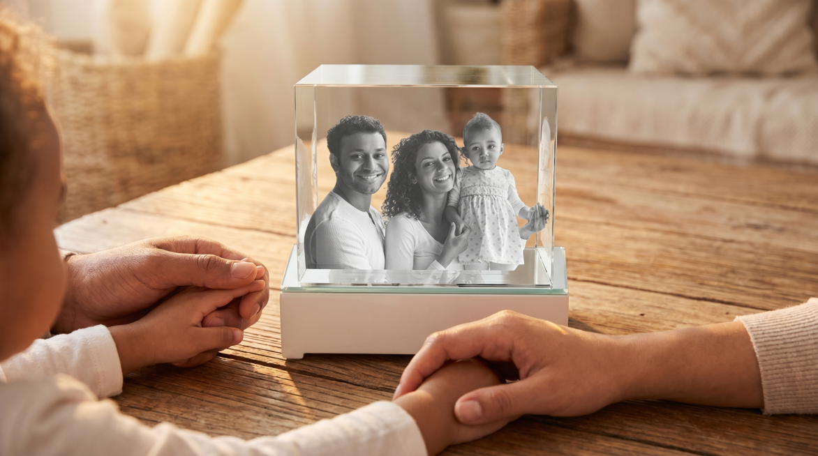 保持原图中玻璃展示盒摆件的外形不变，将摆件放在木质桌面上，桌面伸出年轻爸爸、妈妈和三岁孩子的三只手牵在一起，三只手将摆件包围在中间，营造出温馨温暖的氛围，整体场景美观自然预览效果