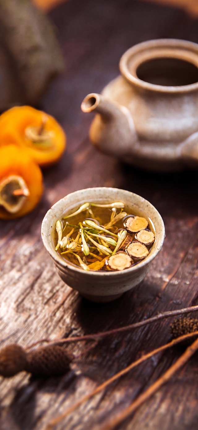 将图中粗陶茶杯内的红豆薏米茶替换为图二中的金银花与甘草组合的花草茶，保留原图的木桌背景、茶壶、柿子等元素及整体暖色调与构图风格