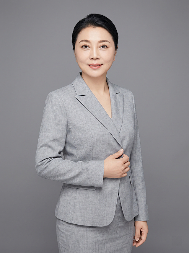 参考图中保留人物脸型、发型、年龄等特征生成职场形象照,标准焦段50mm,f/4光圈,平视视角,镜头与人物视线齐平;人物占画面70%,人物居中,居中构图,位于画面中心,为视觉聚焦点,入境范围头部及以下至臀部下方10cm,半身像;左右留少量背景空间;头部与画面顶部保留5%空间;头部发型完整,头肩比例协调,人物结构准确;发质微微柔顺,微微蓬松。身穿浅灰色精纺羊毛混纺西装外套,平驳领设计,面料呈细腻微微哑光质感;内搭保持原服装风格;下装浅灰色精纺羊毛混纺西装套裙;着装正式干练;专业稳重;身形匀称,姿态端正,身体挺拔。呈右手轻握西装前襟的自然姿态，肢体放松且自信。简约中度灰色纯色背景,呈现均匀柔和质感,背景光线均匀柔和,强化色彩对比的高级感,极简干净;画质超高分辨率、细节清晰,现代精致感风格;肤质精细化处理;眼神直视镜头,目光坚定且澄澈,表情自然。嘴角上扬;清透自然裸妆,轻薄底妆+淡唇妆,妆感轻薄轻透裸肌感,通透感肤质,肤质自然细腻,肤色健康均匀,雾面柔光裸肌感,保留真实皮肤细节。正面柔光补光,4800K中性光,面部光比1:1.5,色调明亮。中近景写实摄影,对焦清晰,高清保留皮肤细节。分辨率4K,去除人物项链配饰，去除人物之外的所有元素。严格按照图片内容生成。画面比例3:4