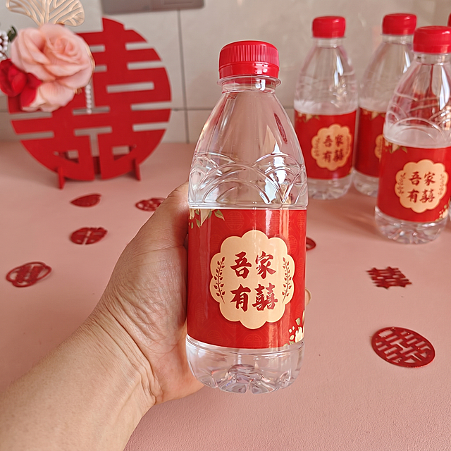 瓶装饮用水，严格参照上传产品图https://gdesign-dam-hz.dancf.com/36462828948014214/16e6c5e5bc804eb59815fa059ce2c00d.png，完整保留所有可见组件、部件、轮廓形状、结构、比例、材质质感、颜色与上传图完全一致，杜绝任何变形、色差、组件缺失、结构偏差。
室内桌面手持拍摄场景，中式婚庆红色暖色调。
一只手手持这瓶饮用水（保留参考图人物性别/国籍/肤色/年龄段/姿态/与产品互动方式），柔和自然光正面侧光（还原参考图自然光方向），产品受光面泛起暖红色、暗部映射浅粉色光晕，与环境物理融合。产品居中特写，微侧 25° 视角。
全量换款清单：①人物：保留原姿态，仅换手型细节与肤色调整，整体相似度50%；②背景道具：参考图大红色喜字剪纸 + 多瓶同款水 + 粉色花艺装饰 → 生成图保留红色喜字元素，调整位置和布局，同品类换款式，背景数量和布局调整保持相似度50%；③桌面承载面：参考图浅粉色平面 → 生成图暖粉色哑光桌面，同材质换纹理细节；④装饰：参考图散落小喜字剪纸 → 生成图同样保留散落中式婚庆剪纸装饰，换具体摆放位置。
去除上传图中的促销文字、价格标签、排版图层等非产品本体元素，仅保留产品本体信息。
4K 分辨率，严格锁定宽高比为 1:1。