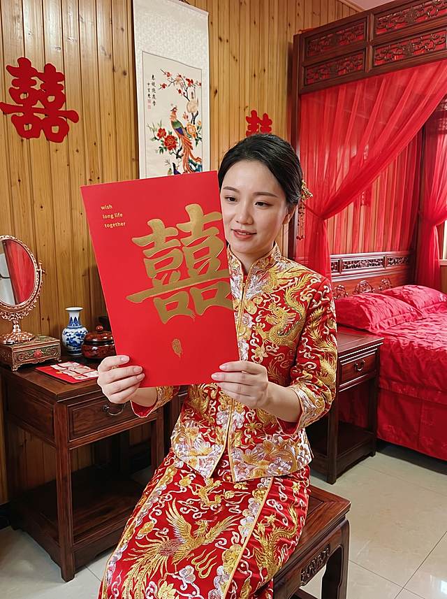 画面中心为身着正红色龙凤褂的新娘，端正坐在闺房的红木雕花凳子上，上半身挺直，轻微向左侧倾斜10°。 红包为左右两页展开式设计，占画面核心视觉区域近 50% 面积，保留原红包样式：右侧页为红色纸币样式，左侧页为烫金大字'囍'红包封面，不做任何改动。背景为复古中式接亲闺房，墙面为浅棕色木质护墙板，悬挂复古中式喜字、刺绣挂画，床头为红木雕花床头，铺着红色提花床品与红色纱幔；床头摆放红木床头柜，上面放着青花瓷花瓶、喜糖罐与中式摆件；一侧摆放复古梳妆台，台上放着胭脂、铜镜与中式首饰盒，光线为暖黄色室内灯，搭配少量自然光，整体氛围典雅复古，充满传统中式韵味，无现代杂乱元素。高清写实手机摄影风格，画面带有生活纪实感，质感清晰细腻，无过度磨皮或美颜，保留真实的皮肤纹理、手部细节、衣物刺绣质感，还原日常随手拍的真实氛围感，光影自然柔和。中近景平视构图，人物居中，红包为绝对视觉焦点，背景轻微虚化突出主体，竖版画面比例，上下留白均衡，视角与日常手机拍摄一致，无夸张透视。