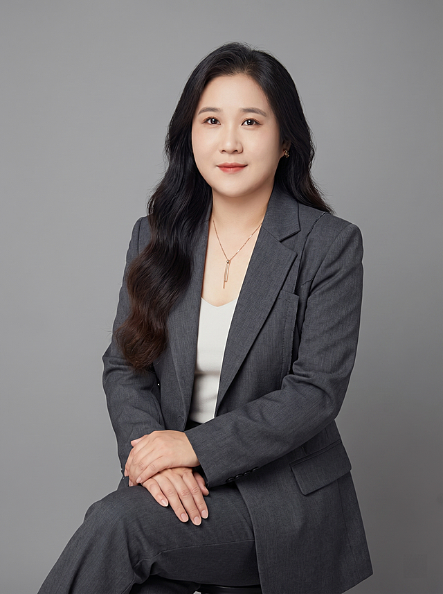 参考图中保留人物脸型、发型、年龄等特征生成职场形象照,标准焦段50mm,f/4光圈(f/2.8-f/4区间),平视视角(镜头与人物视线齐平);人物占画面70%,人物居中(居中构图),位于画面中心,为视觉聚焦点,入境范围头部及以下至臀部下方10cm(半身像);左右留少量背景空间;头部与画面顶部保留5%空间;头部发型完整,头肩比例协调,人物结构准确;身穿深灰色精纺羊毛混纺设计师款西装套装,(解构剪裁设计,面料呈细腻微微哑光质感);内搭简约浅色内搭;下装深灰色精纺羊毛混纺西装下装;着装正式干练;前卫自信,艺术格调;身形匀称,姿态端正,身体挺拔。呈自然坐姿，双手交叠放腿上，肢体放松且自信;发质微微柔顺，微微蓬松。简约微微中度灰色纯色背景(呈现均匀柔和质感),背景光线均匀柔和,强化色彩对比的高级感。极简干净;画质超高分辨率、细节清晰,现代精致感风格;肤质精细化处理;眼神直视镜头,目光坚定且澄澈,表情自然。嘴角上扬;清透自然裸妆(轻薄底妆+淡唇妆)妆感轻薄轻透裸肌感,通透感肤质,肤质自然细腻,肤色健康均匀,雾面柔光裸肌感,保留真实皮肤细节｡正面柔光补光,4800K中性光,面部光比1:1.5,色调明亮｡中近景写实摄影,对焦清晰,高清保留皮肤细节｡佩戴精致配饰，保留原项链和耳环；分辨率4K,去除人物之外的所有元素。严格按照图片内容生成，画面比例3:4