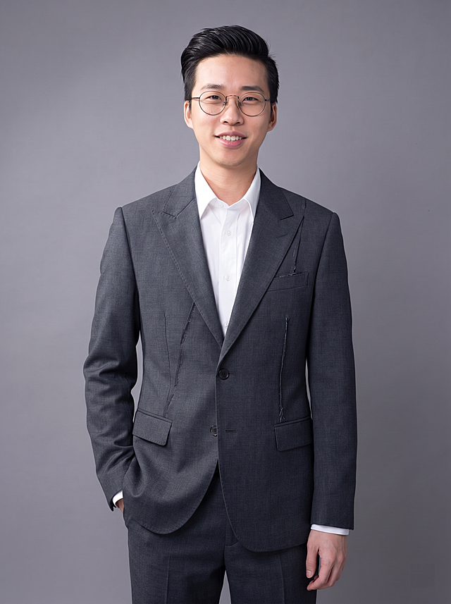 参考图中保留人物脸型、发型、年龄等特征生成设计师职场形象照,标准焦段50mm,f/4光圈(f/2.8-f/4区间),平视视角(镜头与人物视线齐平);人物占画面70%,人物居中(居中构图),位于画面中心,为视觉聚焦点,入境范围头部及以下至臀部下方10cm(半身像);左右留少量背景空间;头部与画面顶部保留5%空间;头部发型完整,头肩比例协调,人物结构准确;发质微微柔顺，微微蓬松。身穿深灰色精纺羊毛混纺设计师款解构剪裁西装外套;内搭白色高支精梳棉衬衫;下装深灰色精纺羊毛混纺西裤;着装正式干练;前卫自信、灵感充沛、艺术格调、张扬有态度;身形匀称,姿态端正,身体挺拔。呈自然站姿，左手插兜，肢体放松且自信;佩戴框架眼镜;简约中性灰纯色背景(呈现均匀柔和质感),背景光线均匀柔和,强化色彩对比的高级感,极简干净;画质超高分辨率、细节清晰,现代精致感风格;肤质精细化处理;眼神直视镜头,目光坚定且澄澈,表情自然。嘴角上扬;肤质自然细腻,肤色健康均匀,皮肤状态清爽干净,保留真实皮肤细节｡正面柔光补光,4800K中性光,面部光比1:1.5｡中近景写实摄影,对焦清晰,面部雾面柔光,轻透裸肌感,高清保留皮肤细节｡分辨率4K,去除人物之外的所有元素。严格按照图片内容生成。画面比例3:4