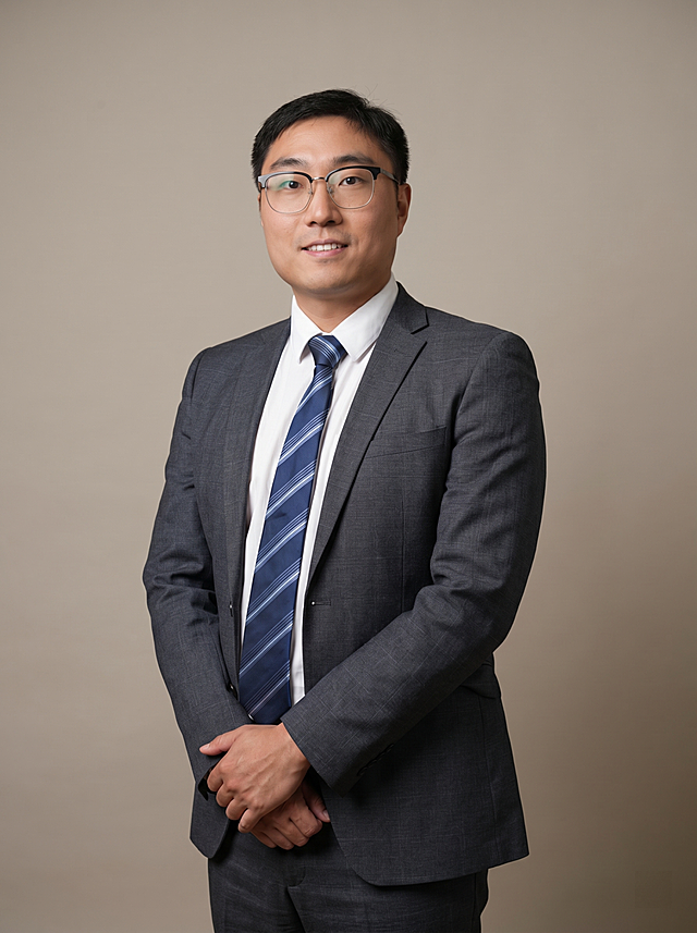 参考图中保留人物脸型、发型、年龄等特征生成金融法律行业职场形象照，标准焦段50mm，f/4光圈，平视视角(镜头与人物视线齐平);人物占画面70%，人物居中(居中构图)，位于画面中心，为视觉聚焦点，入境范围头部及以下至臀部下方10cm(半身像);左右留少量背景空间;头部与画面顶部保留5%空间;头部发型完整，头肩比例协调，人物结构准确;发质微微柔顺，微微蓬松。身穿深灰色精纺羊毛混纺西装外套，(平驳领设计，面料呈细腻微微哑光质感);内搭白色高支精梳棉衬衫;下装深灰色精纺羊毛混纺西裤;系蓝条纹领带，着装正式干练;严谨理性，逻辑清晰，不怒自威，公信力强;身形匀称，姿态端正，身体挺拔。呈自然站姿，双手交叉，肢体放松且自信;佩戴眼镜;简约微微暖调暖灰/米杏纯色背景(呈现均匀柔和质感)，背景光线均匀柔和，强化色彩对比的高级感，极简干净;画质超高分辨率、细节清晰，现代精致感风格;肤质精细化处理;眼神直视镜头，目光坚定且澄澈，表情自然。嘴角上扬;肤质自然细腻，肤色健康均匀，皮肤状态清爽干净，保留真实皮肤细节｡正面柔光补光，4800K中性光，面部光比1:1.5｡中近景写实摄影，对焦清晰，面部雾面柔光，轻透裸肌感，高清保留皮肤细节｡分辨率4K，去除人物之外的所有元素。严格按照图片内容生成。画面比例3:4