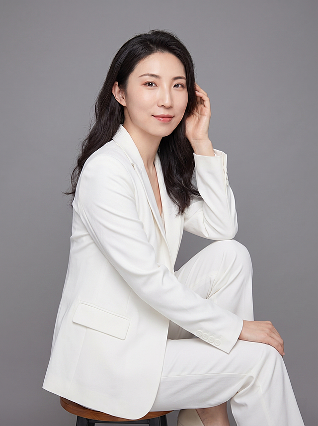 参考图中保留人物脸型、发型、年龄等特征生成休闲个人形象照,标准焦段50mm,f/4光圈(f/2.8-f/4区间),平视视角(镜头与人物视线齐平);人物占画面70%,人物居中(居中构图),位于画面中心,为视觉聚焦点,入境范围头部及以下至臀部下方10cm(半身像);左右留少量背景空间;头部与画面顶部保留5%空间;头部发型完整,头肩比例协调,人物结构准确;发质微微柔顺，微微蓬松。身穿白色休闲西装外套,(平驳领设计,面料细腻微微哑光质感);内搭简约内搭;下装同色系休闲下装;着装休闲干练;自然优雅,轻松自信;身形匀称,姿态端正,身体挺拔。呈侧坐坐姿，单手轻放头部按头的自然姿态，肢体放松且自信;保留原有口红和配饰特征;对脸部做自然简单修饰;简约微微中度灰色纯色背景(呈现均匀柔和质感),背景光线均匀柔和,强化色彩对比的高级感。极简干净;画质超高分辨率、细节清晰,现代精致感风格;肤质精细化处理;眼神直视镜头,目光坚定且澄澈,表情自然。嘴角上扬;清透自然裸妆(轻薄底妆+淡唇妆)妆感轻薄轻透裸肌感,通透感肤质,肤质自然细腻,肤色健康均匀,雾面柔光裸肌感,保留真实皮肤细节｡正面柔光补光,4800K中性光,面部光比1:1.5,色调明亮｡中近景写实摄影,对焦清晰,高清保留皮肤细节｡分辨率4K,去除人物之外的所有元素。严格按照图片内容生成。画面比例3:4