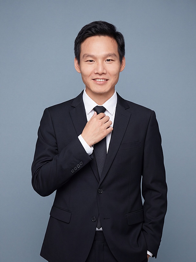 参考图中保留人物脸型、发型、年龄等特征生成教育/学术领域职场形象照,标准焦段50mm,f/4光圈(f/2.8-f/4区间),平视视角(镜头与人物视线齐平);人物占画面70%,人物居中(居中构图),位于画面中心,为视觉聚焦点,入境范围头部及以下至臀部下方10cm(半身像);左右留少量背景空间;头部与画面顶部保留5%空间;头部发型完整,头肩比例协调,人物结构准确;发质微微柔顺，微微蓬松。身穿深黑色精纺羊毛混纺西装外套,(平驳领设计,面料呈细腻微微哑光质感);内搭白色高支精梳棉衬衫;下装深黑色精纺羊毛混纺西裤;着装正式干练;博学温润,学者风范,从容儒雅,知性内敛;身形匀称,姿态端正,身体挺拔。呈手扶领带的自然站姿，肢体放松且自信;简约冷灰蓝灰色纯色背景(呈现均匀柔和质感),背景光线均匀柔和,强化色彩对比的高级感,极简干净;画质超高分辨率、细节清晰,现代精致感风格;肤质精细化处理;眼神直视镜头,目光坚定且澄澈,表情自然。嘴角上扬;肤质自然细腻,肤色健康均匀,皮肤状态清爽干净,保留真实皮肤细节｡正面柔光补光,4800K中性光,面部光比1:1.5｡中近景写实摄影,对焦清晰,面部雾面柔光,轻透裸肌感,高清保留皮肤细节｡分辨率4K,去除人物之外的所有元素。严格按照图片内容生成。画面比例3:4