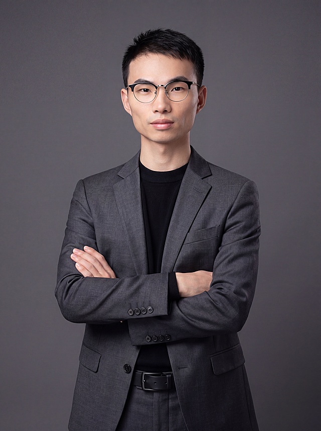 参考图中保留人物脸型、发型、年龄等特征生成职场形象照,标准焦段50mm,f/4光圈(f/2.8-f/4区间),平视视角(镜头与人物视线齐平);人物占画面70%,人物居中(居中构图),位于画面中心,为视觉聚焦点,入境范围头部及以下至臀部下方10cm(半身像);左右留少量背景空间;头部与画面顶部保留5%空间;头部发型完整,头肩比例协调,人物结构准确;发质微微柔顺，微微蓬松。身穿修身精纺羊毛混纺面料的深灰色西装外套(含5%桑蚕丝提升柔光质感),平驳领(商务感正式感),左胸袋无口袋巾(简化高级感),西装下摆自然垂度;内搭黑色中领紧身针织上衣,下装精纺羊毛混纺面料的深灰色高腰直筒西裤(面料同西装),搭配黑色哑光头层牛皮皮带(宽1.5cm),带扣为长方形拉丝黑色金属(1.8cm×2.5cm);着装正式干练;正式严谨专业稳重;身形匀称,姿态端正,身体挺拔。呈正面抱胸的自然站立姿态:左臂搭于右臂上方,肩颈自然松弛下沉,姿态自信从容;佩戴圆形细框眼镜;画质超高分辨率、细节清晰,现代精致感风格;肤质精细化处理;眼神直视镜头,目光坚定且澄澈,表情自然。简约冷调深灰色纯色背景(呈现均匀柔和质感),背景光线均匀柔和,强化色彩对比的高级感，极简干净;肤质自然细腻,肤色健康均匀,皮肤状态清爽干净,保留真实皮肤细节｡正面柔光补光,4800K中性光,面部光比1:1.5｡中近景写实摄影,对焦清晰,面部雾面柔光,轻透裸肌感,高清保留皮肤细节。分辨率4K,去除人物之外的所有元素。严格按照图片内容生成。画面比例3:4