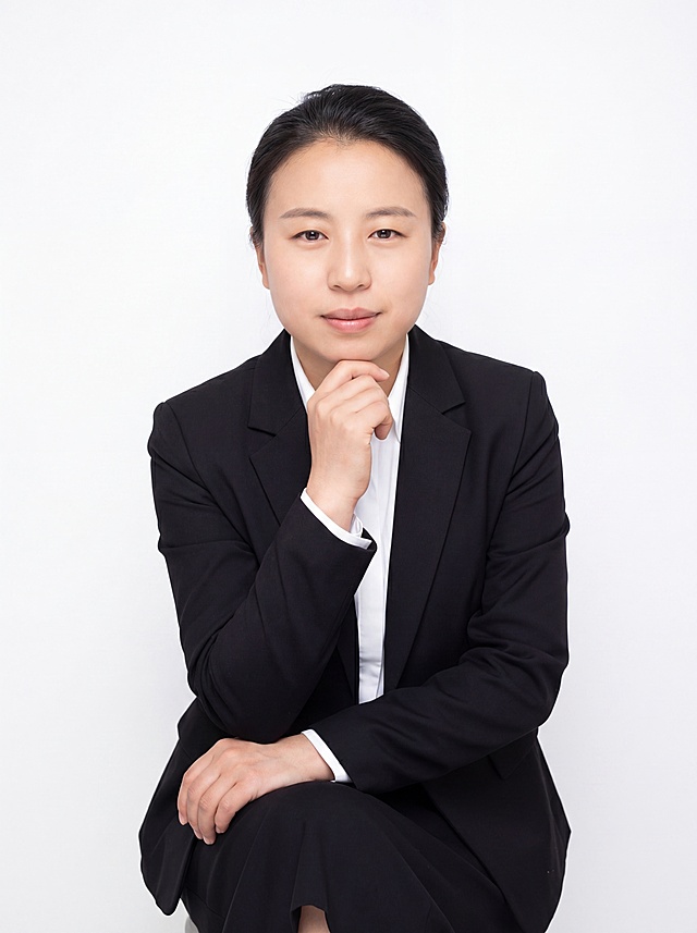 参考图中保留人物脸型、发型、年龄等特征生成职场形象照,标准焦段50mm,f/4光圈(f/2.8-f/4区间),平视视角(镜头与人物视线齐平);人物占画面70%,人物居中(居中构图),位于画面中心,为视觉聚焦点,入境范围头部及以下至臀部下方10cm(半身像);左右留少量背景空间;头部与画面顶部保留5%空间;头部发型完整,头肩比例协调,人物结构准确;发质微微柔顺，微微蓬松。身穿黑色精纺羊毛混纺西装外套,(平驳领设计,面料呈细腻微微哑光质感);内搭白色高支精梳棉衬衫;下装黑色精纺羊毛混纺西装套裙;着装正式干练;专业稳重;身形匀称,姿态端正,身体挺拔。呈坐姿，手扶下巴的自然姿态，肢体放松且自信;简约纯白色纯色背景(呈现均匀柔和质感),背景光线均匀柔和,强化色彩对比的高级感。极简干净;画质超高分辨率、细节清晰,现代精致感风格;肤质精细化处理;眼神直视镜头,目光坚定且澄澈,表情自然。嘴角上扬;清透自然裸妆(轻薄底妆+淡唇妆)妆感轻薄轻透裸肌感,通透感肤质,肤质自然细腻,肤色健康均匀,雾面柔光裸肌感,保留真实皮肤细节｡正面柔光补光,4800K中性光,面部光比1:1.5,色调明亮｡中近景写实摄影,对焦清晰,高清保留皮肤细节｡分辨率4K,去除人物之外的所有配饰，去除项链/耳环、包包、帽子等配饰。严格按照图片内容生成。画面比例3:4