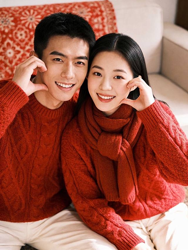 两人身穿红色粗线针织毛衣、米白色下装，女生搭配同色围巾，两人亲密依偎，同时用单手在脸颊旁比出爱心手势，两人笑容灿烂，轻松自然的姿势，温馨居家室内环境，背景是红色花纹软装、浅色调沙发，光线柔和温暖，中近景构图，平视角度，胶片质感情侣写真
