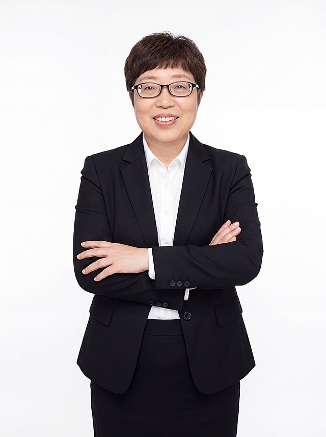 参考图中保留人物脸型、发型、年龄等特征生成职场形象照,标准焦段50mm,f/4光圈(f/2.8-f/4区间),平视视角(镜头与人物视线齐平);人物占画面70%,人物居中(居中构图),位于画面中心,为视觉聚焦点,入境范围头部及以下至臀部下方10cm(半身像);左右留少量背景空间;头部与画面顶部保留5%空间;头部发型完整,头肩比例协调,人物结构准确;发质微微柔顺，微微蓬松。身穿黑色精纺羊毛混纺西装外套,(平驳领设计,面料呈细腻微微哑光质感);内搭白色高支精梳棉衬衫;下装黑色精纺羊毛混纺西装套裙;着装正式干练;沉稳可靠、专业公正;身形匀称,姿态端正,身体挺拔。呈站姿，双手抱胸，肢体放松且自信;保留眼镜配饰，去除细项链及其他配饰;简约纯白色纯色背景(呈现均匀柔和质感),背景光线均匀柔和,强化色彩对比的高级感。极简干净;画质超高分辨率、细节清晰,现代精致感风格;肤质精细化处理;眼神直视镜头,目光坚定且澄澈,表情自然。嘴角上扬;清透自然裸妆(轻薄底妆+淡唇妆)妆感轻薄轻透裸肌感,通透感肤质,肤质自然细腻,肤色健康均匀,雾面柔光裸肌感,保留真实皮肤细节｡正面柔光补光,4800K中性光,面部光比1:1.5,色调明亮｡中近景写实摄影,对焦清晰,高清保留皮肤细节;分辨率4K,去除人物之外原背景和细项链等多余配饰，只保留眼镜。严格按照图片内容生成。画面比例3:4