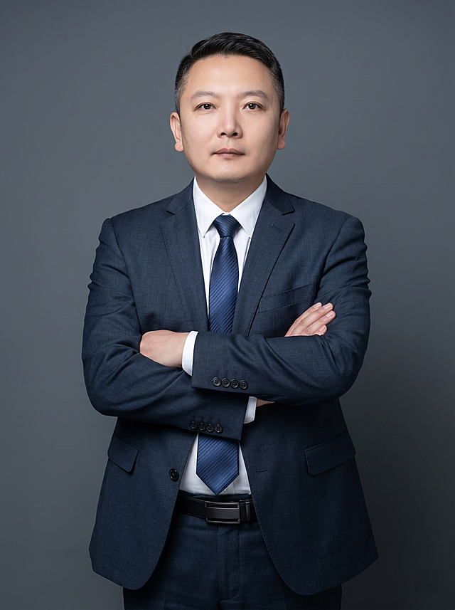 参考图中保留人物脸型、发型、年龄等特征生成职场形象照，标准焦段50mm,F/4光圈(处于F/2.8-F/4区间),平视视角(镜头与人物视线齐平);人物占画面70%,人物居中(居中构图),位于画面中心,为视觉聚焦点,入境范围头部及以下至臀部下方10cm(半身像)构图，左右留少量背景空间;头部与画面顶部保留5%空间;头部发型完整,头肩比例协调,人物结构准确;身穿精纺羊毛混纺面料的深墨蓝色西装外套(含5%桑蚕丝提升柔光质感),平驳领(商务感正式感),左胸袋无口袋巾(简化高级感),西装下摆自然垂度;内搭长绒棉白色衬衫(温莎领),袖口贴合手腕1cm(无堆叠);搭配精致且高级的深蓝色斜向微微渐隐光泽感极细条纹商务感领带(丝提花材质,低饱和柔光渐隐);下装精纺羊毛混纺面料的深墨蓝色高腰直筒西裤(面料同西装);搭配黑色哑光头层牛皮皮带(宽2.5cm),带扣为长方形拉丝黑色金属(1.8cm×3cm);着装正式干练;专业稳重，身形匀称,姿态端正,身体挺拔。呈正面抱胸的自然站立姿态:左臂搭于右臂上方,肩颈自然松弛下沉,姿态自信从容，正式严谨专业稳重;发梢微微修整,干练大气,主体锐利,简约冷调深灰色纯色背景(呈现均匀柔和质感),背景光线均匀柔和,强化色彩对比的高级感，极简干净背景;画质超高分辨率、细节清晰,现代精致感风格;肤质精细化处理;眼神直视镜头,目光坚定且澄澈,表情自然。肤质自然细腻,肤色健康均匀,哑光肤质清爽干净,保留真实皮肤细节;杰作,最佳画质,超高细节,写实摄影,中近景人像,皮肤质感自然清晰,柔光,正面补光,画面干净清新,高清皮肤细节;面部光比1:1.5,4800K中性光,微微修饰脸型,肤质哑光细腻,面部雾面柔光,轻透裸肌感,真实毛孔纹理;分辨率4K,严格按照图片内容生成，画面比例3:4